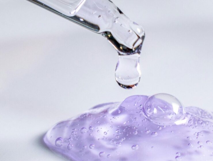 Clear hair serum dripping from a glass dropper onto translucent purple gel.
