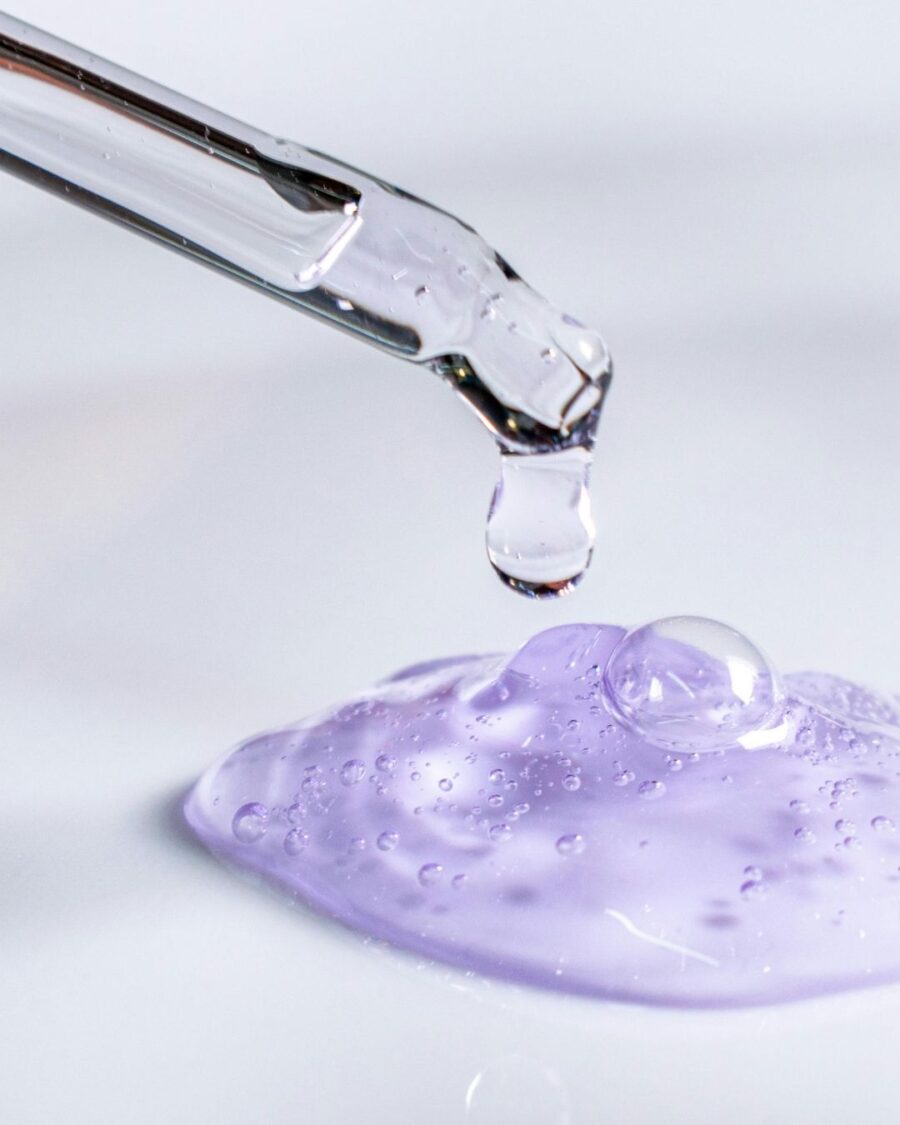 Clear hair serum dripping from a glass dropper onto translucent purple gel.