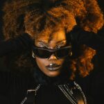 Woman posing with black sunglasses and a black outfit. Her hands are pushing the sides of her hair back.
