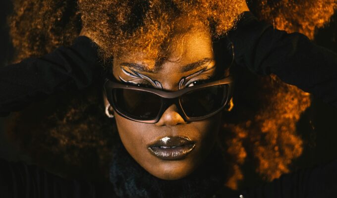 Woman posing with black sunglasses and a black outfit. Her hands are pushing the sides of her hair back.