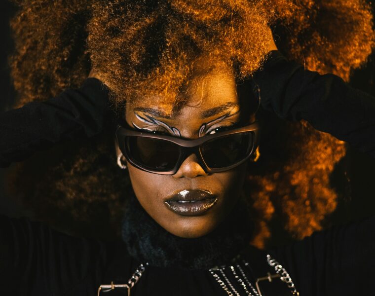 Woman posing with black sunglasses and a black outfit. Her hands are pushing the sides of her hair back.
